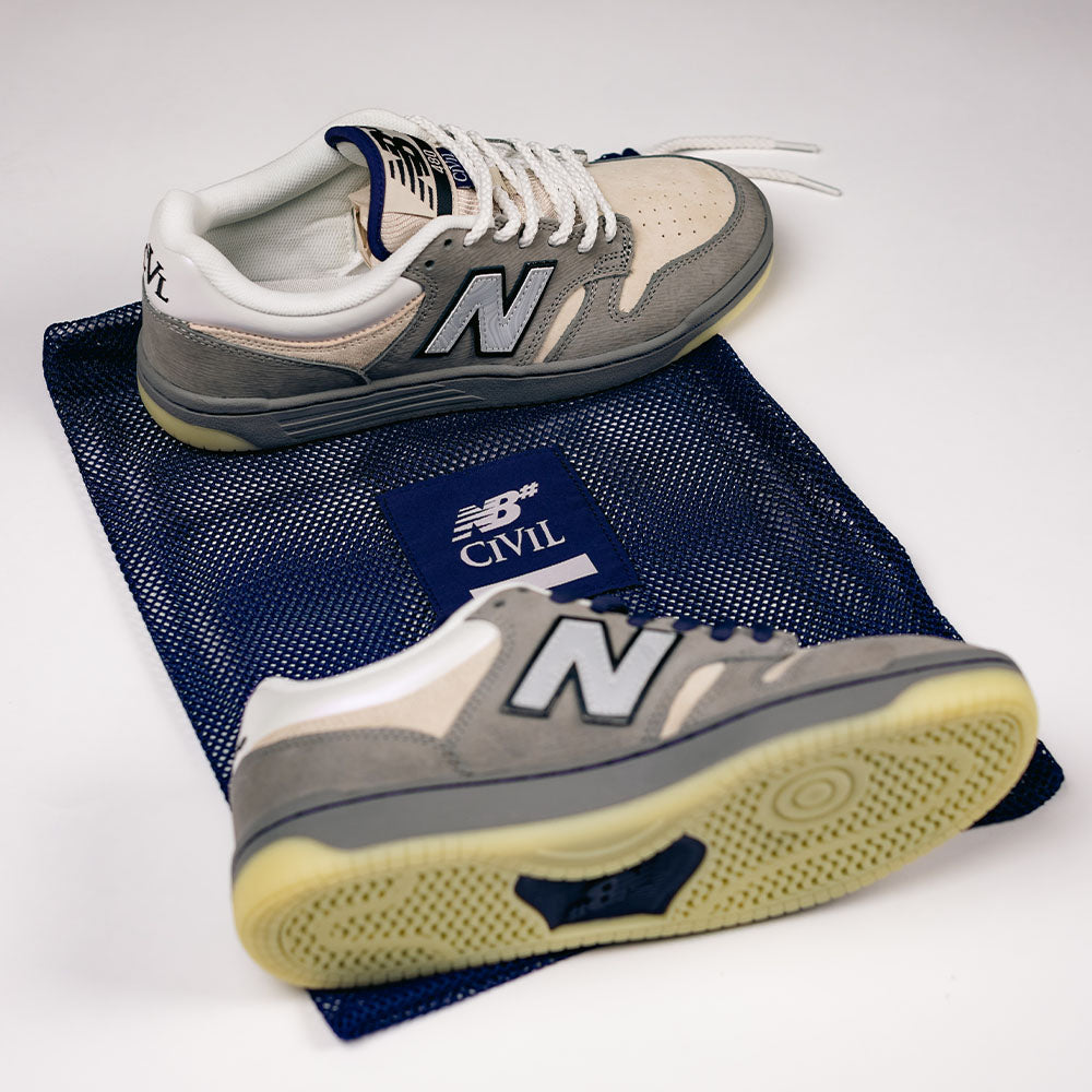 A pair of 480 CVL New Balance Numeric x Civil Suede Leather Skateboarding Shoes in gray and beige rests atop a blue mesh bag marked with the New Balance and Civil logos. One shoe lies flat as the other is tilted to showcase the durable ABZORB midsole on the sole.