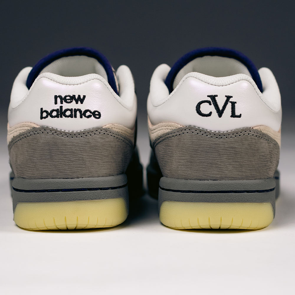 A close-up of the back of two 480 CVL New Balance Numeric x Civil Suede Leather Skateboarding Shoes shows the left shoe with “New Balance” text and the right with “CVL” text. These durable skate shoes are crafted in shades of gray, white, and cream with yellowed soles, and feature an ABZORB midsole for enhanced comfort.