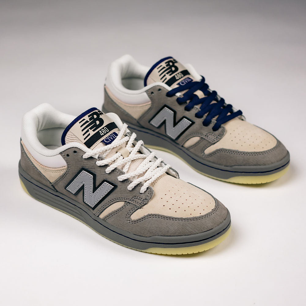 A pair of 480 CVL New Balance Numeric x Civil Suede Leather Skateboarding Shoes in beige, gray, and white—with mismatched laces (one white, one navy blue)—set against a plain light background. Designed for durability, these skate shoes feature an ABZORB midsole to provide superior cushioning and support.