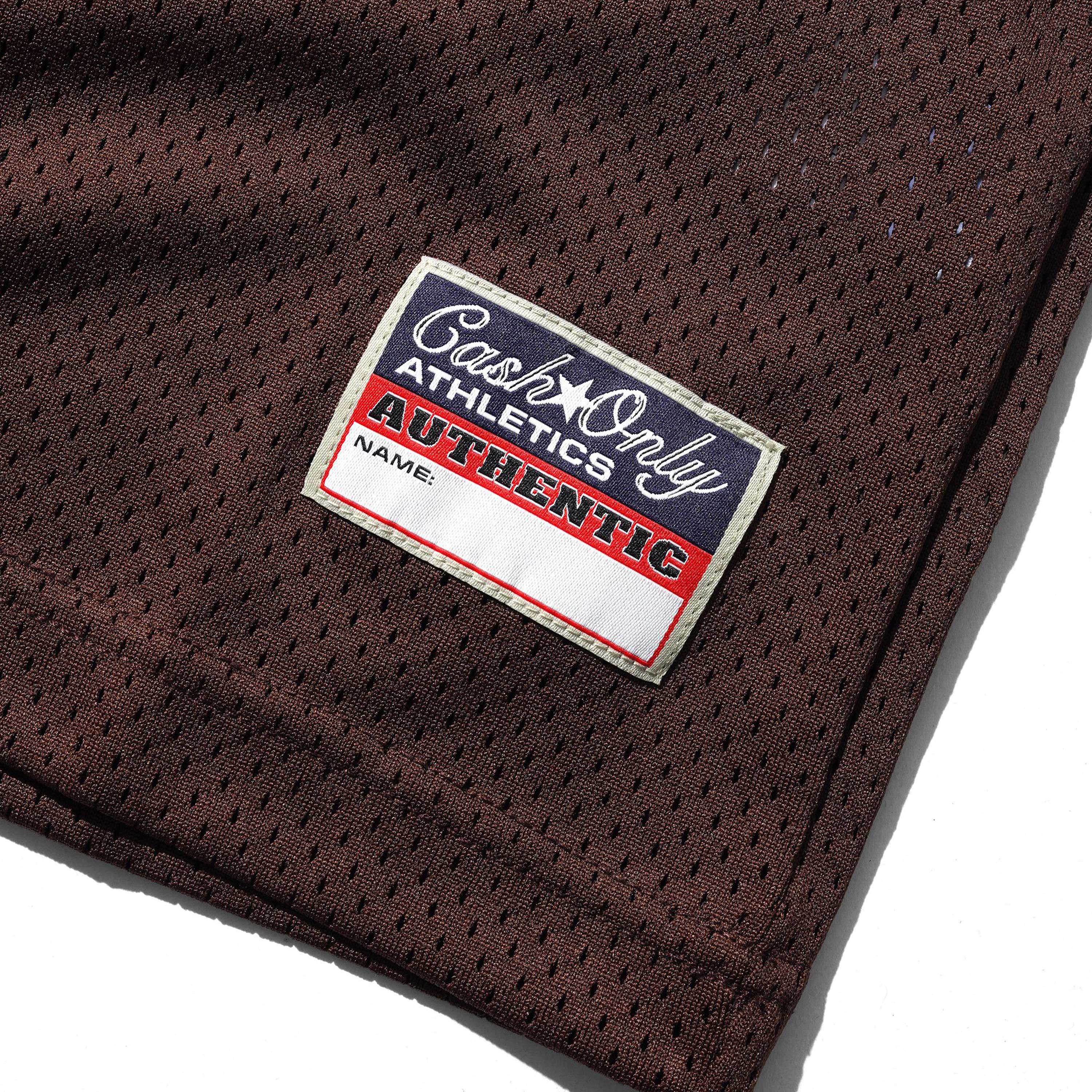 A close-up of the Colt Jersey Brown Cash Only shows its brown polyester fabric and a woven label reading “Cash Only ATHLETICS AUTHENTIC” with a blank line marked “NAME.”.