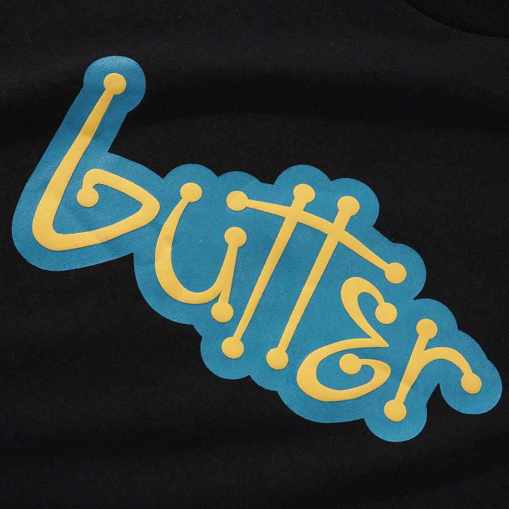 The Connection Black Tee by Butter Goods features the word "butter" in a playful yellow font with dotted ends, set against a blue cloud-like background.