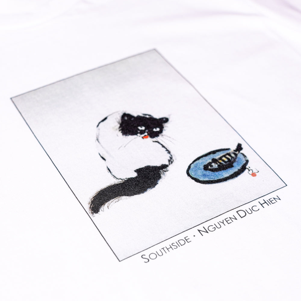 The Curious Cat Southside White Tee by Nguyen Duc Hien features a minimalist illustration of a black and white cat gazing at a blue plate with a fish skeleton, printed on premium USA cotton. “SOUTHSIDE - NGUYEN DUC HIEN” is printed below.