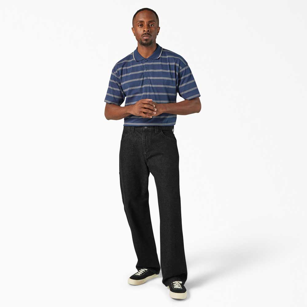 A man stands against a white background, wearing a blue and gray striped polo, DDSK68 Loose Fit Denim Pant Carpenter Black Dickies Skateboarding jeans, and black-and-white sneakers, facing forward with his hands clasped.