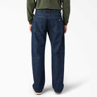 A person wearing DDSK68 Loose Fit Denim Pant Carpenter Dark Wash Dickies Skateboarding jeans and a long-sleeve green shirt stands facing away, showcasing the back pockets of the relaxed fit jeans against a plain white background.