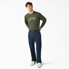 A young man stands against a white background in a green long-sleeve shirt, DDSK68 Loose Fit Denim Pant Carpenter Dark Wash by Dickies Skateboarding, and white sneakers. He has short, tousled hair and poses casually with his hands in his pockets.
