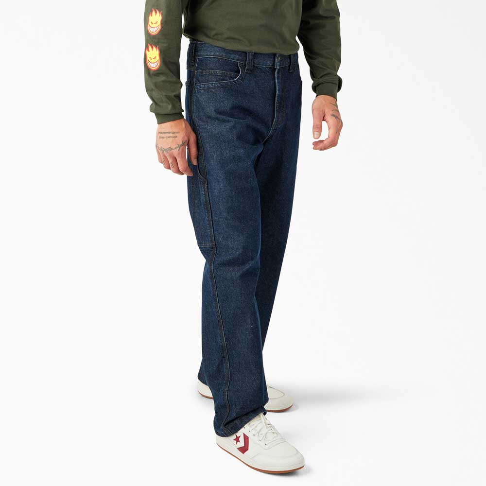 A person wearing DDSK68 Loose Fit Denim Pant Carpenter Dark Wash Dickies Skateboarding jeans, a green long-sleeve shirt with a flame sleeve design, and white sneakers with red accents stands against a plain white background.