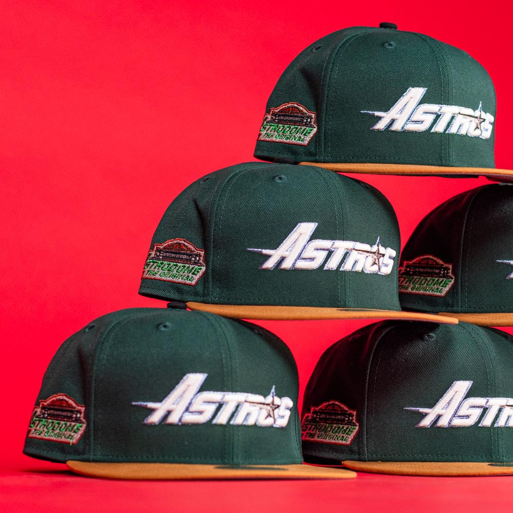 Five El Tuga Houston Astros New Era 5950 Fitted hats in dark green with orange brims are stacked against a bright red background. Each features the Astros logo and a stadium patch on the side.
