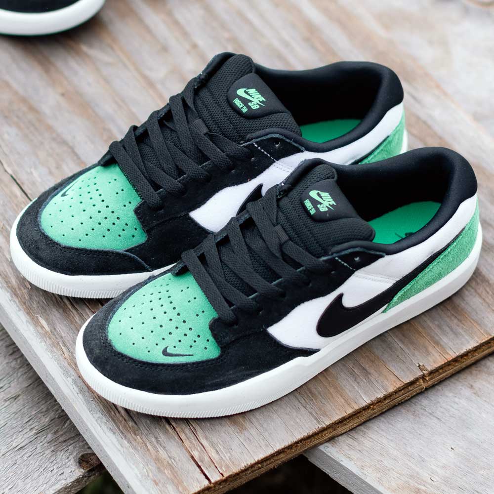 A pair of DV5477101 White Black Stadium Green Force 58 Nike SB suede skateboarding shoes with green perforated toes, black laces, black and white panels, and green insoles sit on a wooden surface.