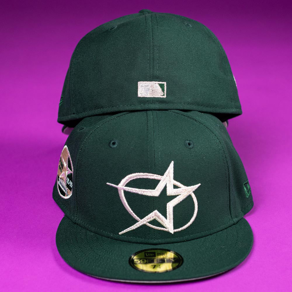 Two The Merc Green Silver 35th Houston Astros New Era Hat 5950 Fitted caps in dark green rest on a purple surface; the front displays a white star and oval, while the back shows a silver MLB logo above the closure.