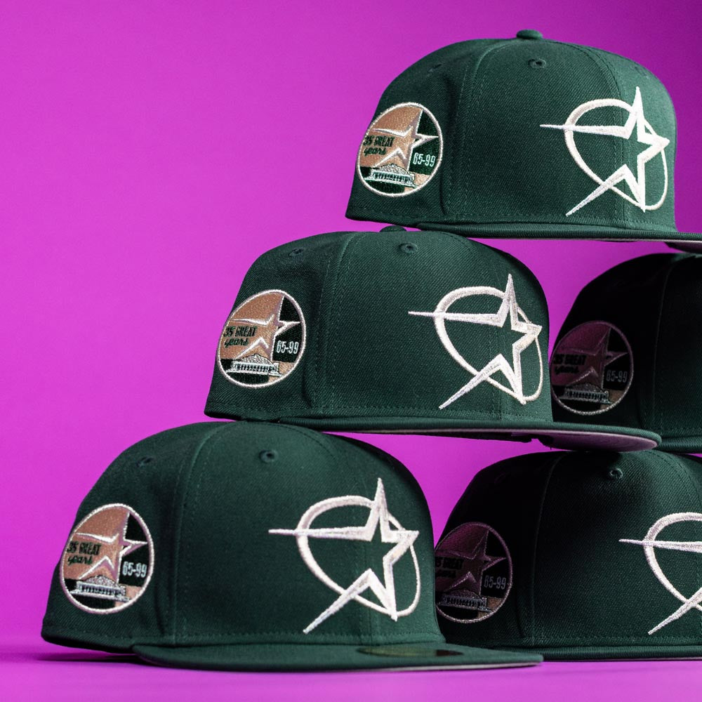 A stack of The Merc Green Silver 35th Houston Astros New Era Hat 5950 Fitted caps featuring a white star logo and circular patch is arranged against a vibrant purple background.