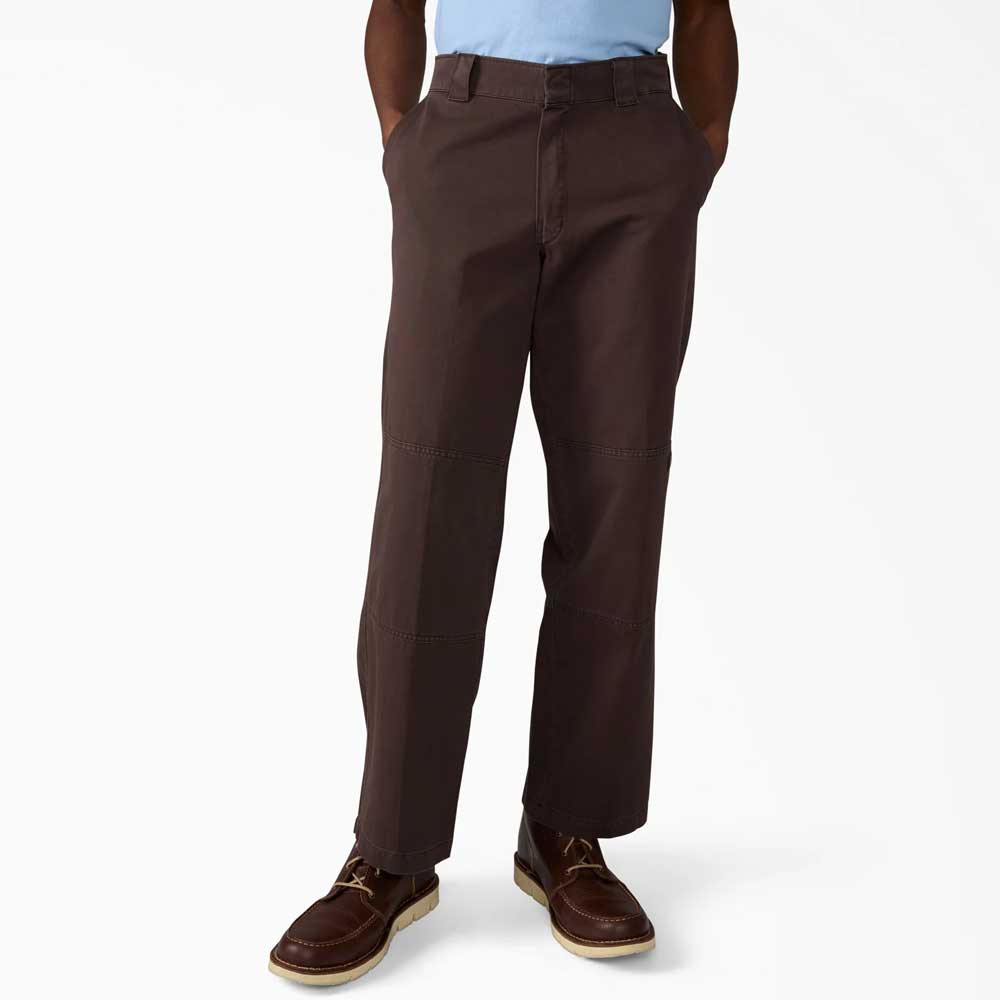 A person wearing Dark Brown Worn In Double Knee Pant A89TC0DB Dickies Skateboarding, a light blue shirt, and brown lace-up shoes stands with hands in pockets against a plain white background.