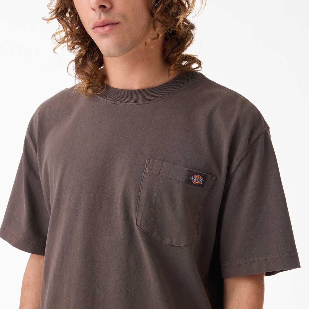 A person with long curly hair wears a relaxed-fit Dark Brown Worn In Heavyweight Pocket Tee A89G70DB by Dickies Skateboarding, featuring a chest pocket with small logo patch. The 100% cotton shirt is cropped at the mouth and mid-arm in the image.