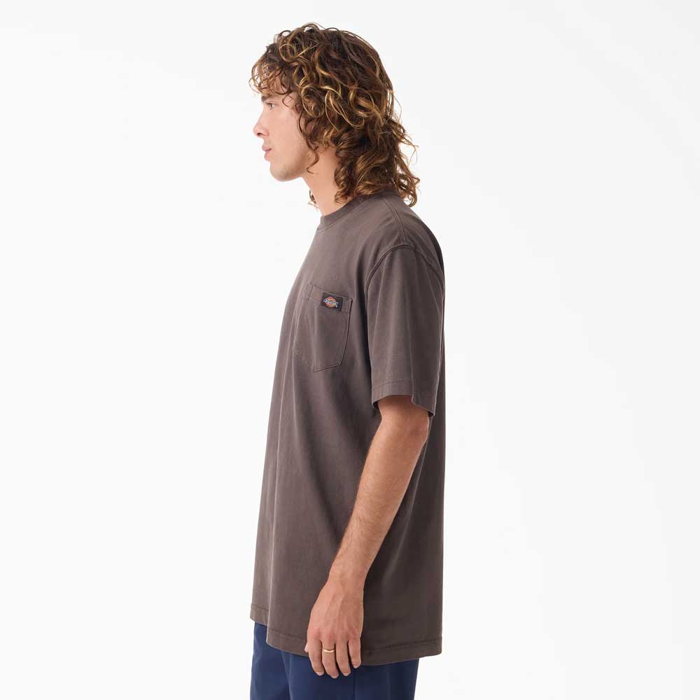 A man with curly hair stands in profile wearing the Dark Brown Worn In Heavyweight Pocket Tee A89G70DB Dickies Skateboarding— a relaxed fit, 100% cotton T-shirt with a chest pocket— and dark pants against a plain white background.