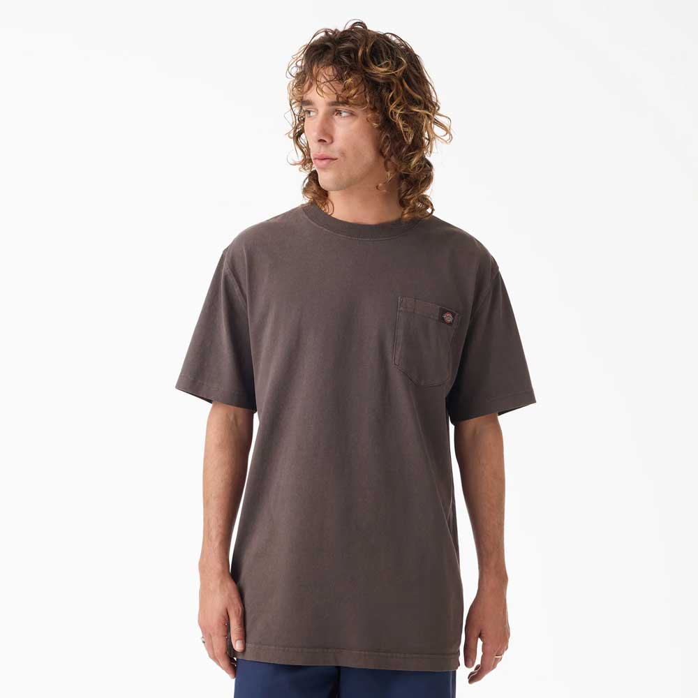 A person with curly, shoulder-length hair wears the Dark Brown Worn In Heavyweight Pocket Tee A89G70DB Dickies Skateboarding and dark blue pants, standing against a plain white background and looking slightly to the side.
