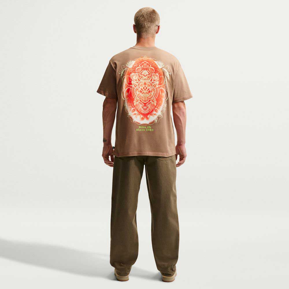A person faces away, wearing the Nike SB Dark Driftwood Som Tum Dunk Tee IH1478258— a roomy midweight cotton T-shirt with a bold red and orange back graphic—paired with loose brown pants and beige shoes against a plain light background.
