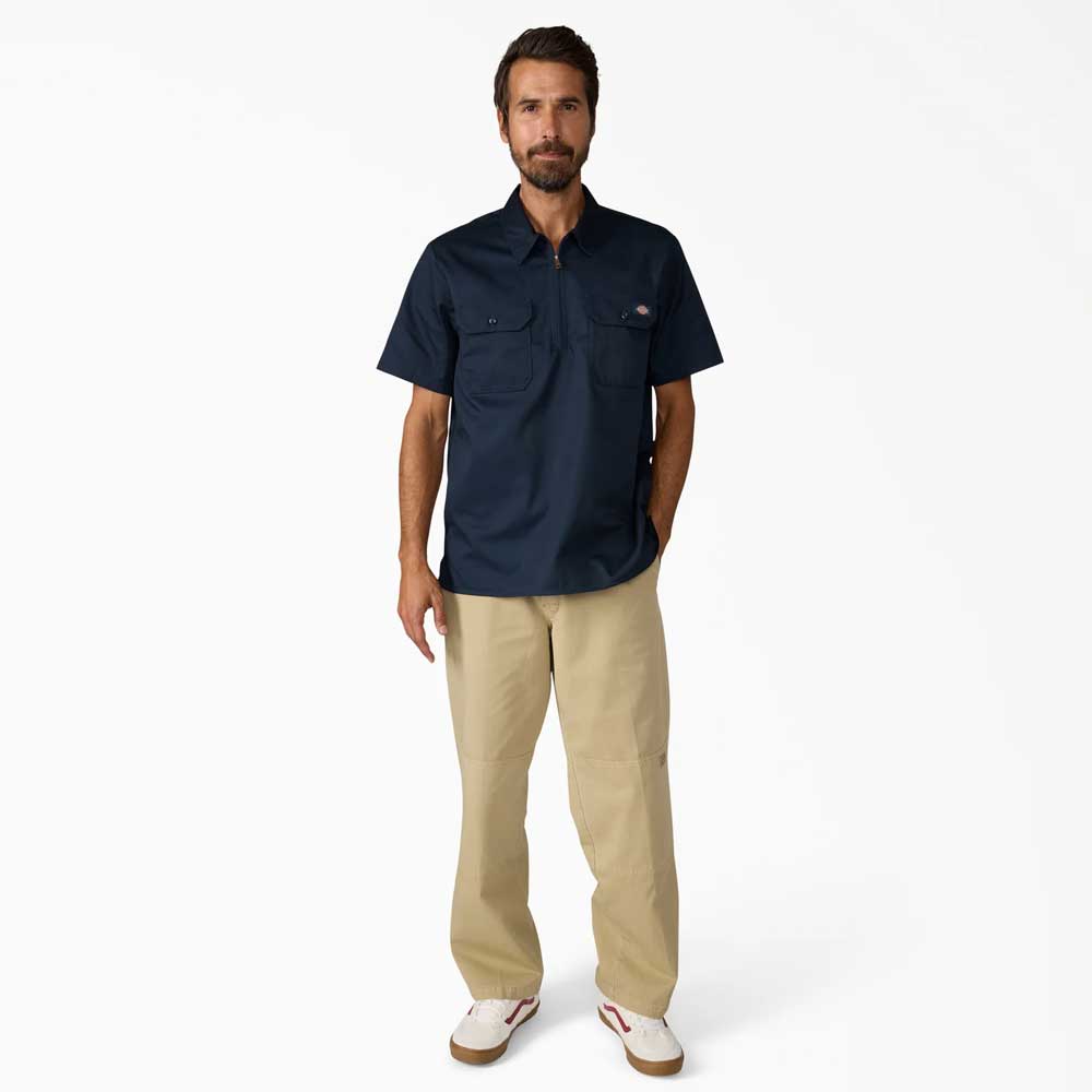 A man faces forward wearing the Dickies Skateboarding Dark Navy 1/4 Zip Short Sleeve Work Shirt A88A10DN, beige pants, and white sneakers with red accents, set against a plain white background.