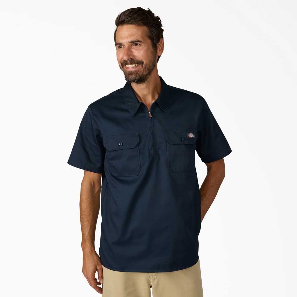 A man with dark hair and a beard stands smiling against a white background, wearing tan pants and a Dickies Skateboarding Dark Navy 1/4 Zip Short Sleeve Work Shirt (A88A10DN) with two chest pockets.