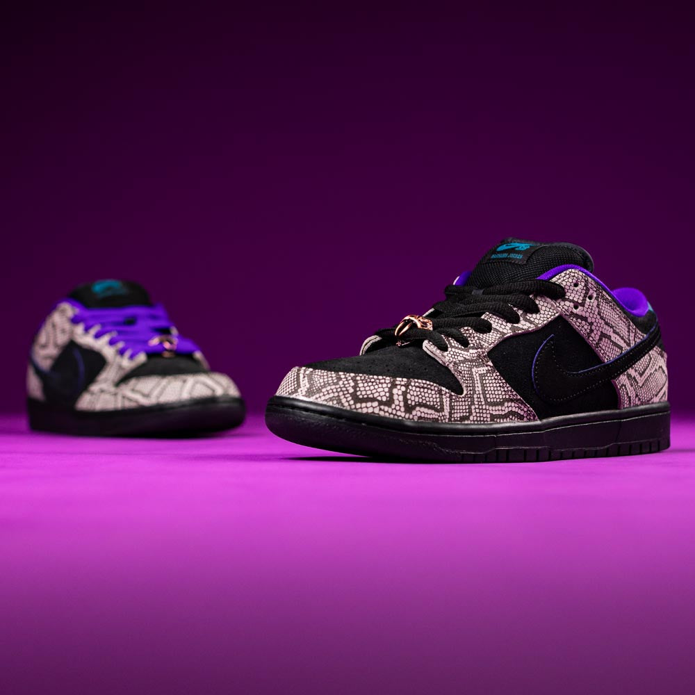 A pair of READ BELOW the BUTTON RAFFLE Nike SB Dashawn Jordan Dunk Low sneakers in black and purple with a grey patterned overlay are shown on a purple surface, inspired by Southside Skatepark. One shoe is in focus, the other blurred.