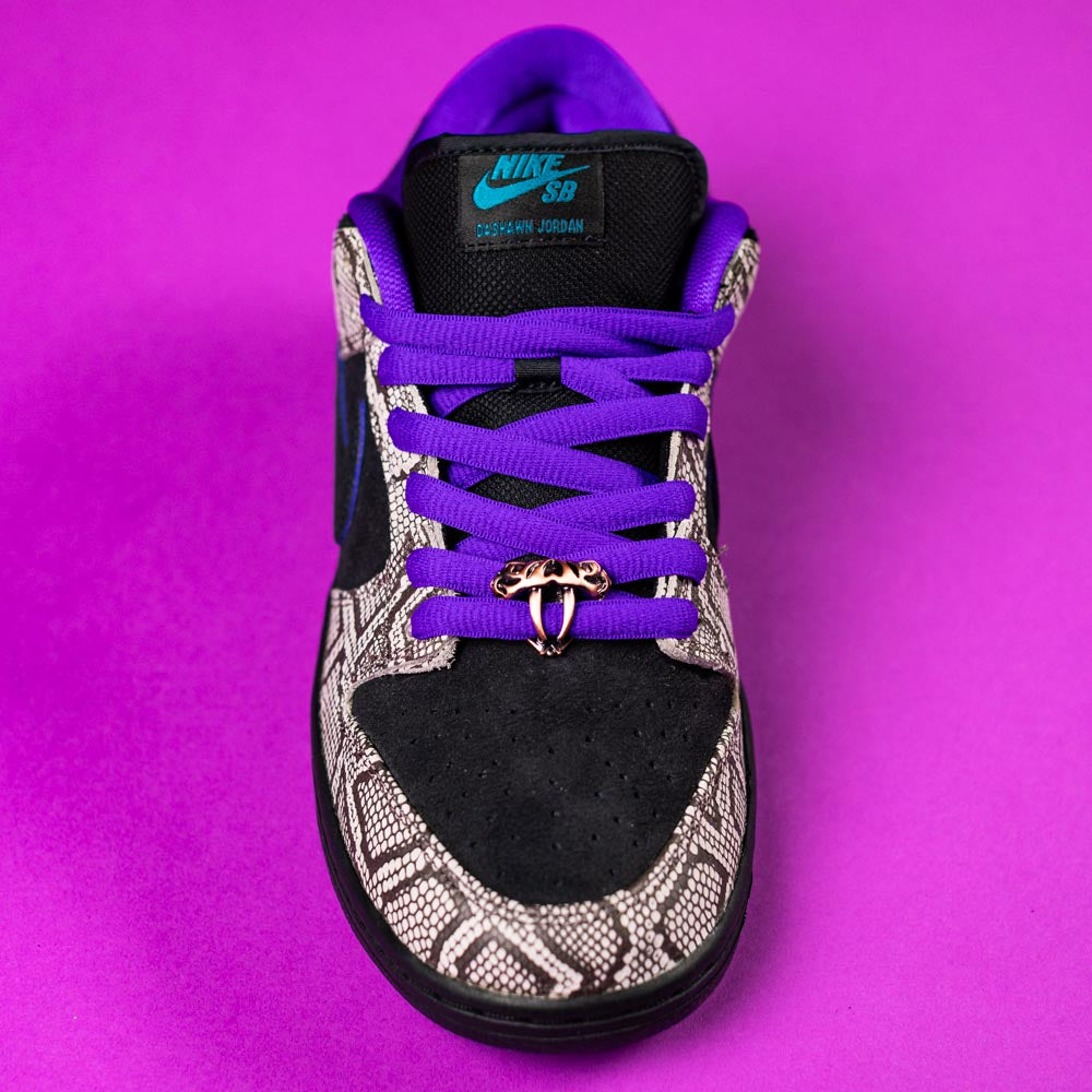 The READ BELOW the BUTTON RAFFLE Nike SB Dashawn Jordan Dunk Low, with black suede, grey snakeskin accents, purple laces, and a gold ring lace accessory, is showcased on a purple background—ideal for Southside Skatepark's raffle entry.