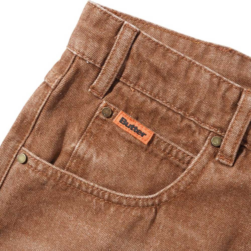Close-up of Distressed Brown Relaxed Denim Jeans by Butter Goods, made from 100% cotton with a small orange "Butter" tag above the pocket, brass rivets at the corners, and visible stitching details.