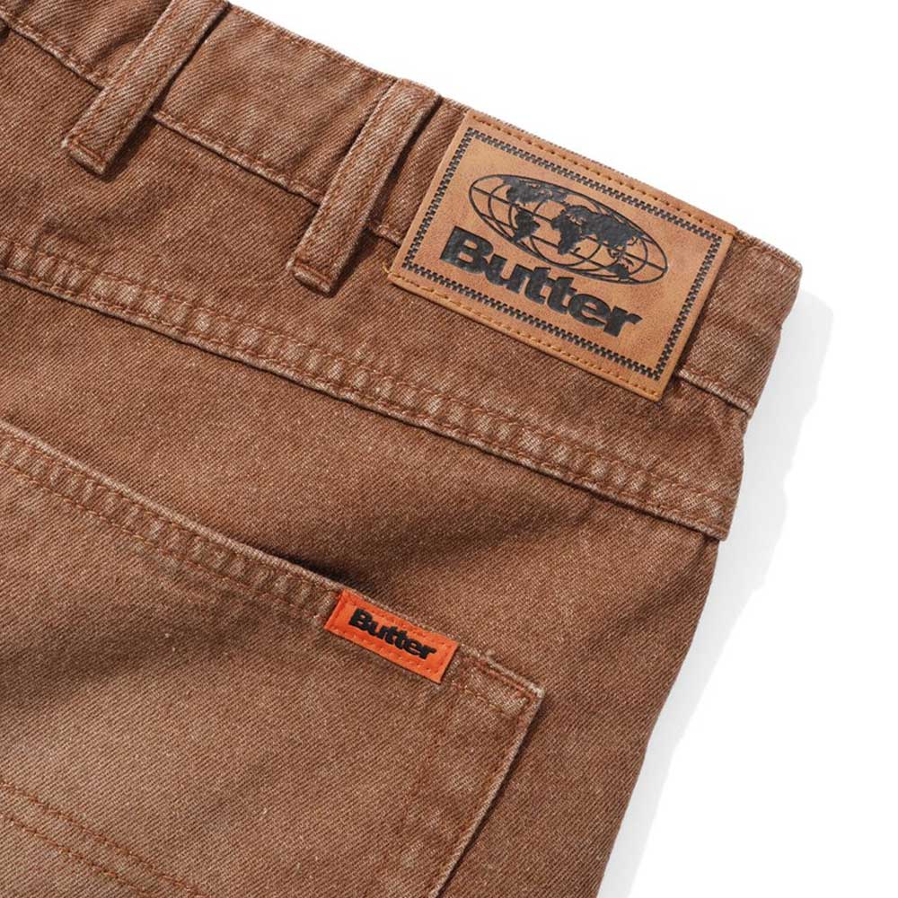 Close-up of the Distressed Brown Relaxed Denim Jeans by Butter Goods, showing a back pocket with a small orange tag and a large leather Butter logo patch above the waistband. Made from 100% cotton denim.