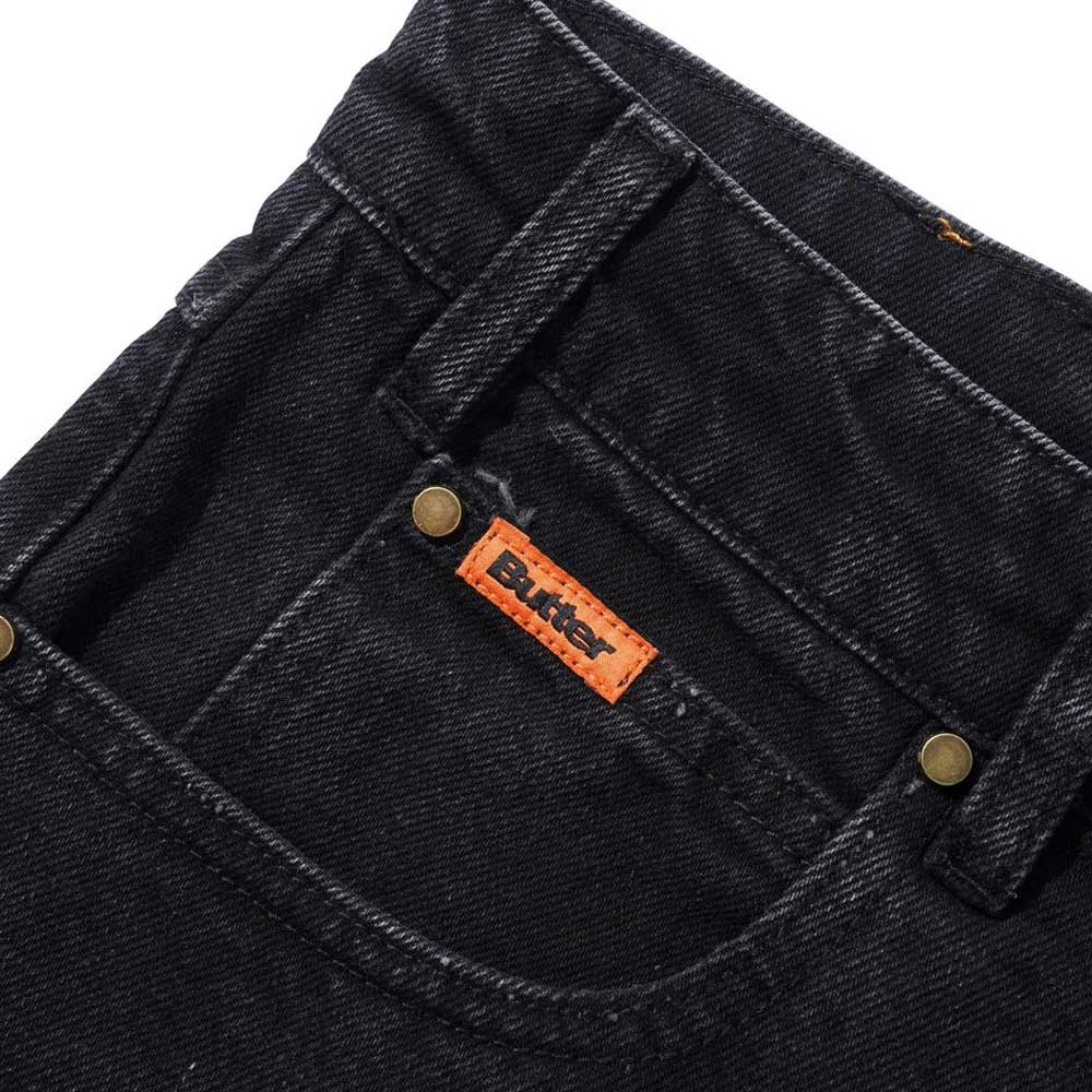 Close-up of the front pocket of Distressed Flat Black Relaxed Denim Jeans by Butter Goods, made from 100% cotton denim with brass rivets, an orange Butter label, and a relaxed fit silhouette.