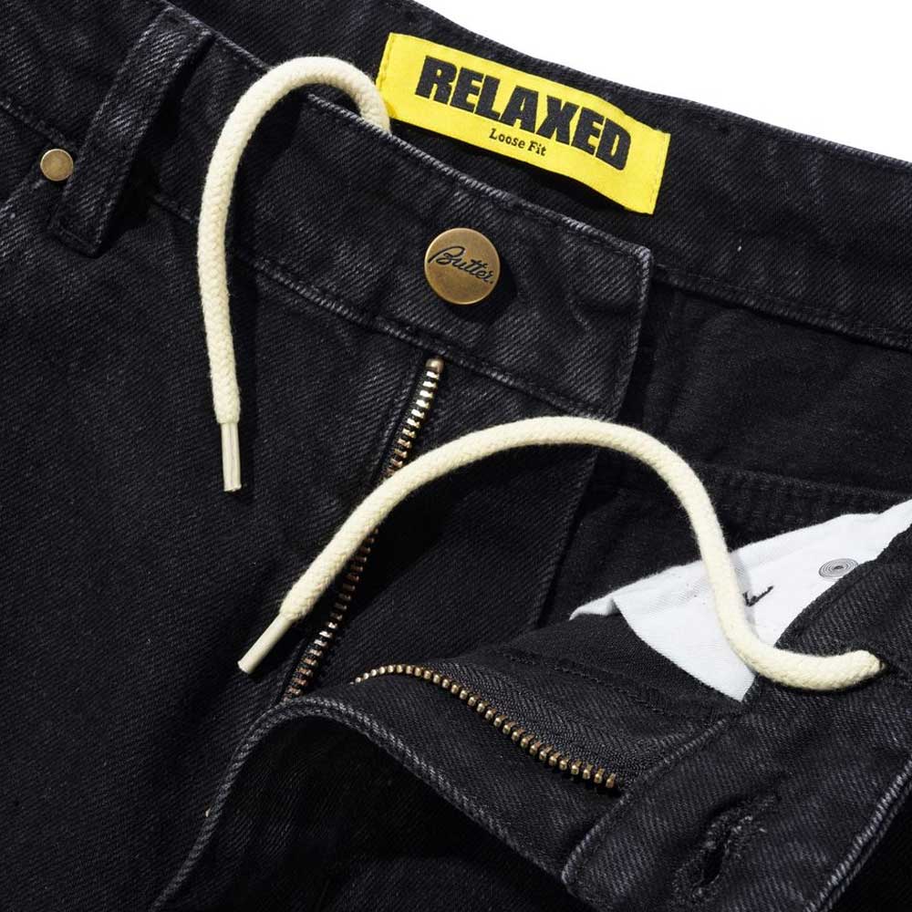 Close-up of Distressed Flat Black Relaxed Denim Jeans by Butter Goods, featuring a white drawstring, visible zipper, yellow RELAXED Loose Fit label, and a button with "Butter" branding above the zipper.
