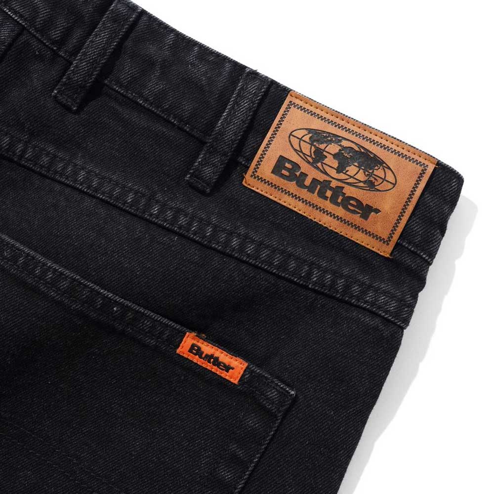 Close-up of Distressed Flat Black Relaxed Denim Jeans by Butter Goods, made from 100% cotton, featuring a brown leather Butter patch with globe graphic and an orange Butter tag above the front pocket.