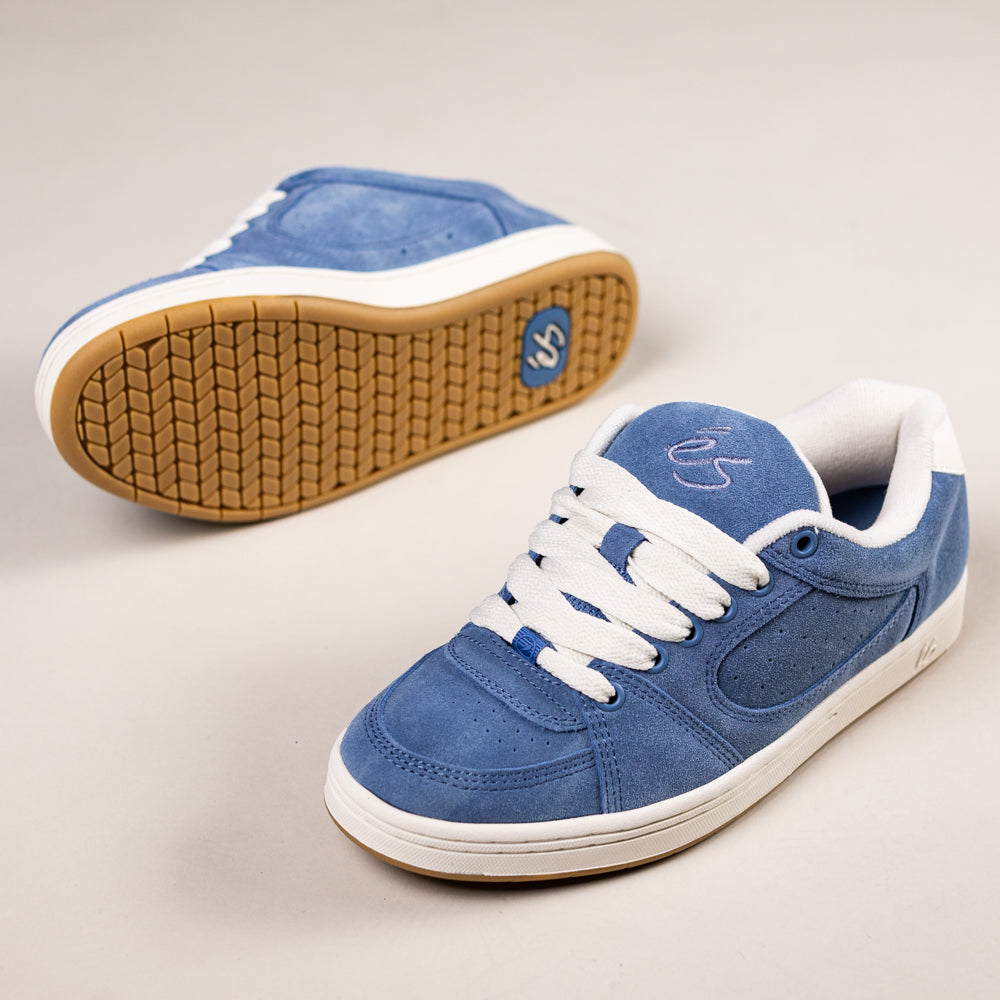 A pair of Accel Blue White Suede Es Skateboarding Shoes is shown—one upright with white laces and sole, the other on its side displaying the gum rubber logo sole. Both rest on a light-colored surface.