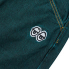 The Eyes Green Overdye Denim Krooked Pant features green overdye denim with yellow stitching and a signature white embroidered patch of two serious cartoon eyes near the pocket.