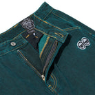 Close-up of Eyes Green Overdye Denim Krooked Pant featuring dark teal-green denim, yellow stitching, metal button, zipper fly, a small embroidered white face on the pocket, and a Krooked pant graphic label sewn inside the waistband.