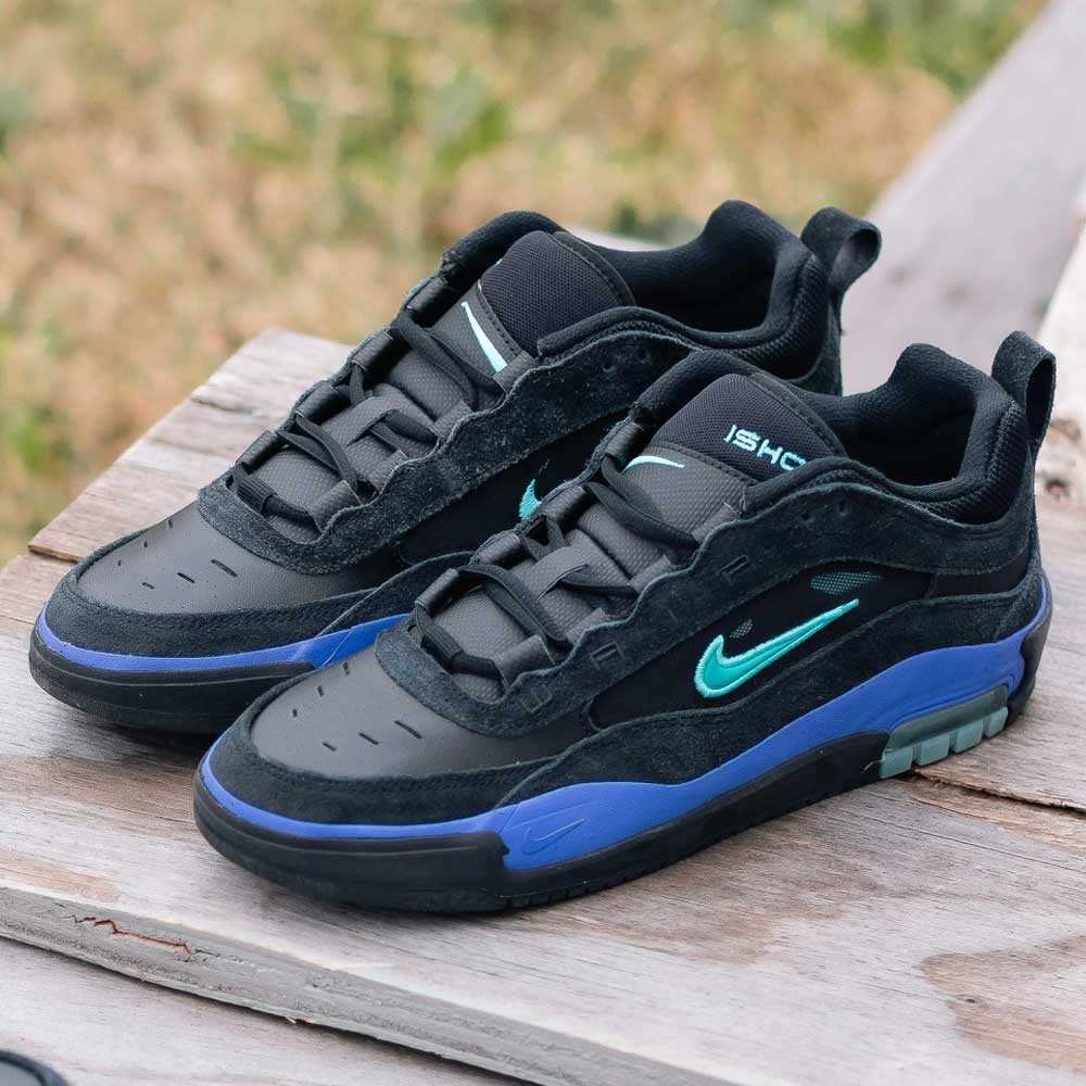 A pair of FB2393-003 Dusty Cactus Black Air Max Ishod Wair Leather Suede Shoes with teal swooshes and blue accents sits on a wooden surface outdoors, grass visible in the background.