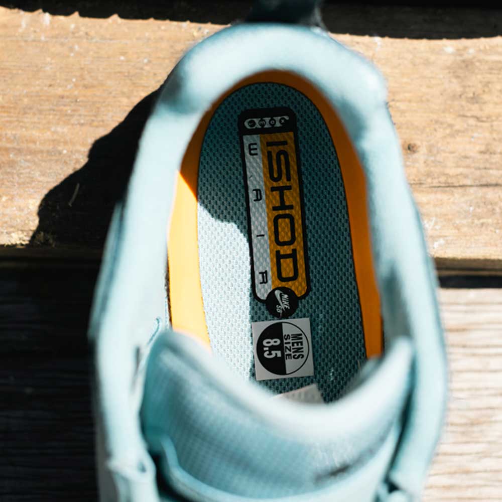 Overhead view of the FB2393400 Denim Turq Black Denim Air MAX Ishod Nike SB Suede Leather Skateboarding Shoe, showing its light blue insole with ISHOD and NIKE SB text, plus a Mens 8.5 size label on a wooden surface.