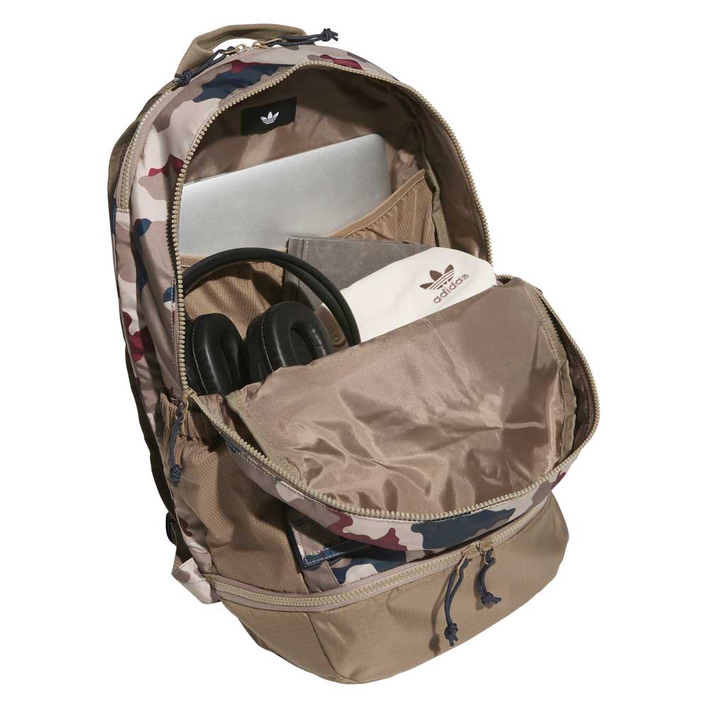 A FSBS Backpack KS6815 Khaki Camo Adidas Skateboarding is shown with its front pocket open, revealing a pair of black headphones, a laptop, and an Adidas pouch inside.
