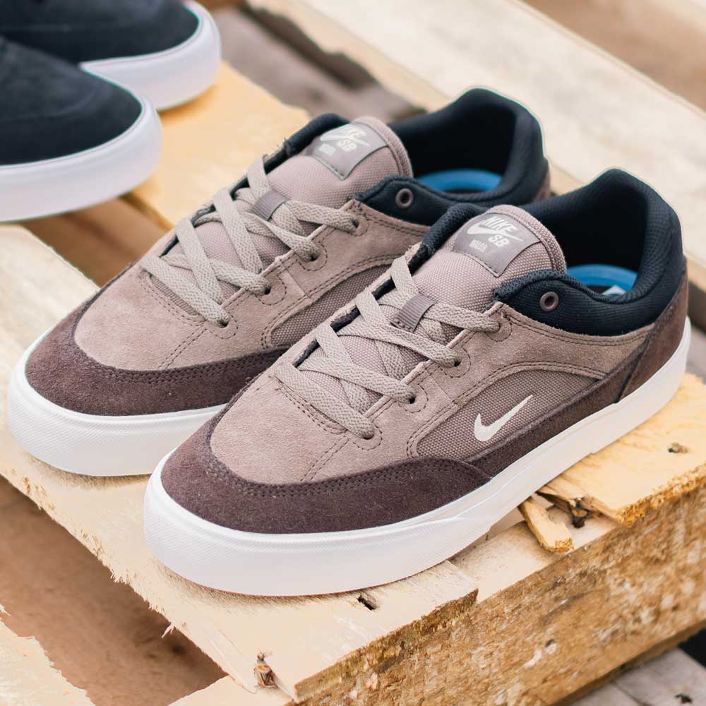 A pair of FV6064005 Ironstone Light Khaki Malor Nike SB suede skateboarding shoes with taupe and brown uppers, white soles, a white Nike swoosh, and black interior lining are displayed on a wooden pallet.