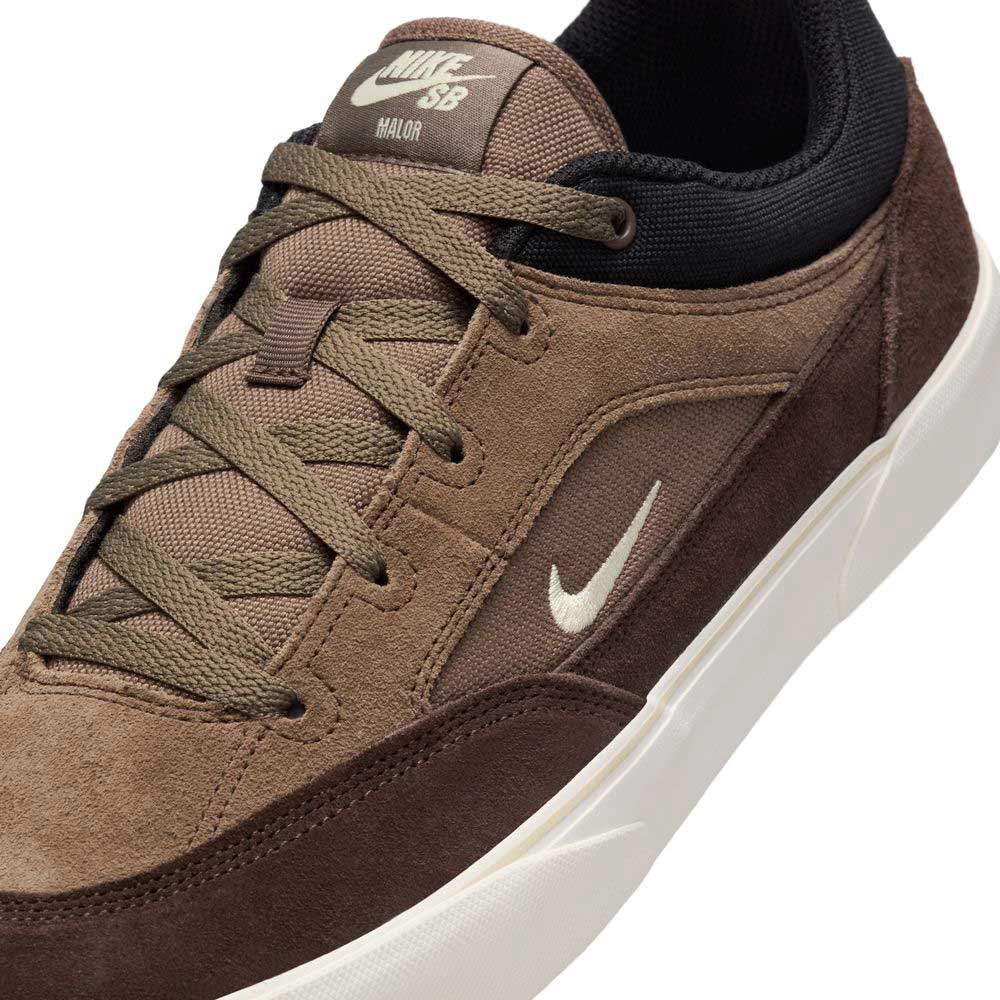 Close-up of the Nike SB Malor FV6064005 Ironstone Light Khaki skateboarding shoes, featuring a brown suede upper, olive green laces, white swoosh and sole, black lining, and the Nike SB logo on the tongue for a stylish finish.