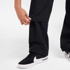 A person wearing the FV7349010 Double Knee Pant Black from Nike SB Skateboarding adjusts the hem of one pant leg with their hand. The background is plain white.