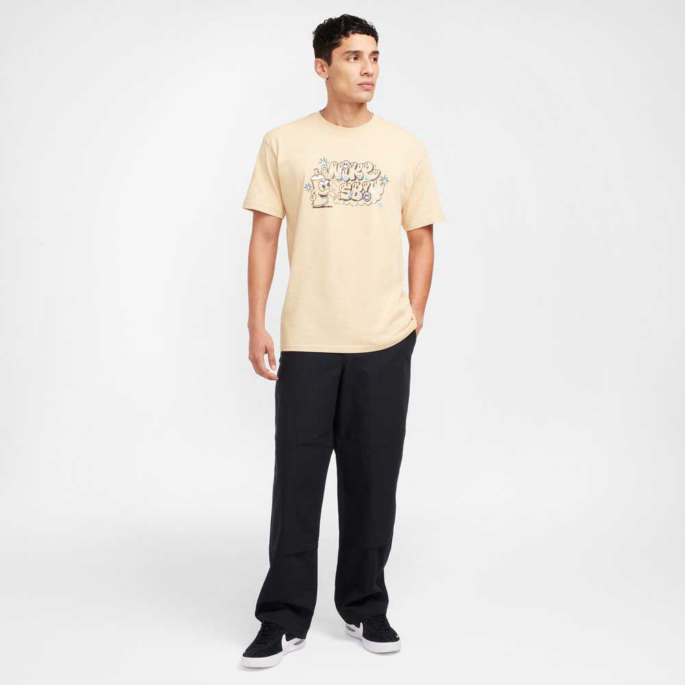A man stands slightly turned, wearing a beige graphic tee, FV7349010 Double Knee Pant Black Nike SB Skateboarding, and black sneakers against a plain white background.