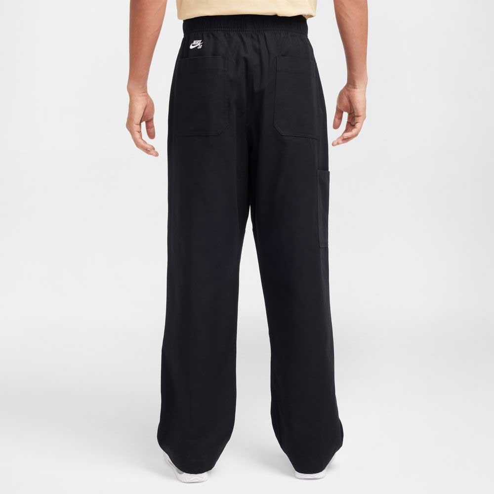 A person faces away, wearing Nike SB Skateboarding FV7349-010 Double Knee Pant in black. The loose-fit cotton twill pants feature patch pockets and a small white Nike logo near the waist, standing out against a light background.