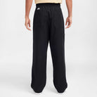 A person faces away, wearing Nike SB Skateboarding FV7349-010 Double Knee Pant in black. The loose-fit cotton twill pants feature patch pockets and a small white Nike logo near the waist, standing out against a light background.