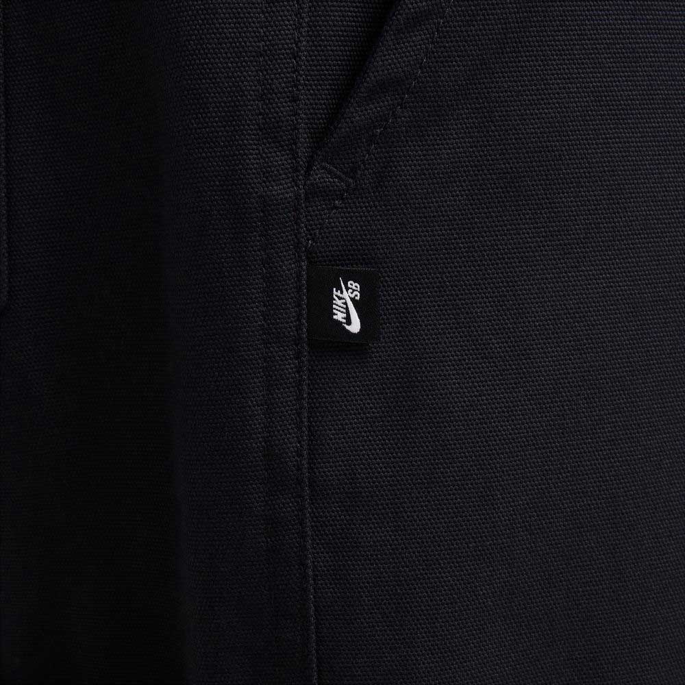 Close-up of black cotton twill fabric from the FV7349010 Double Knee Pant Black Nike SB Skateboarding, showing a small black tag with the white Nike SB logo near a seam, likely on a pocket.