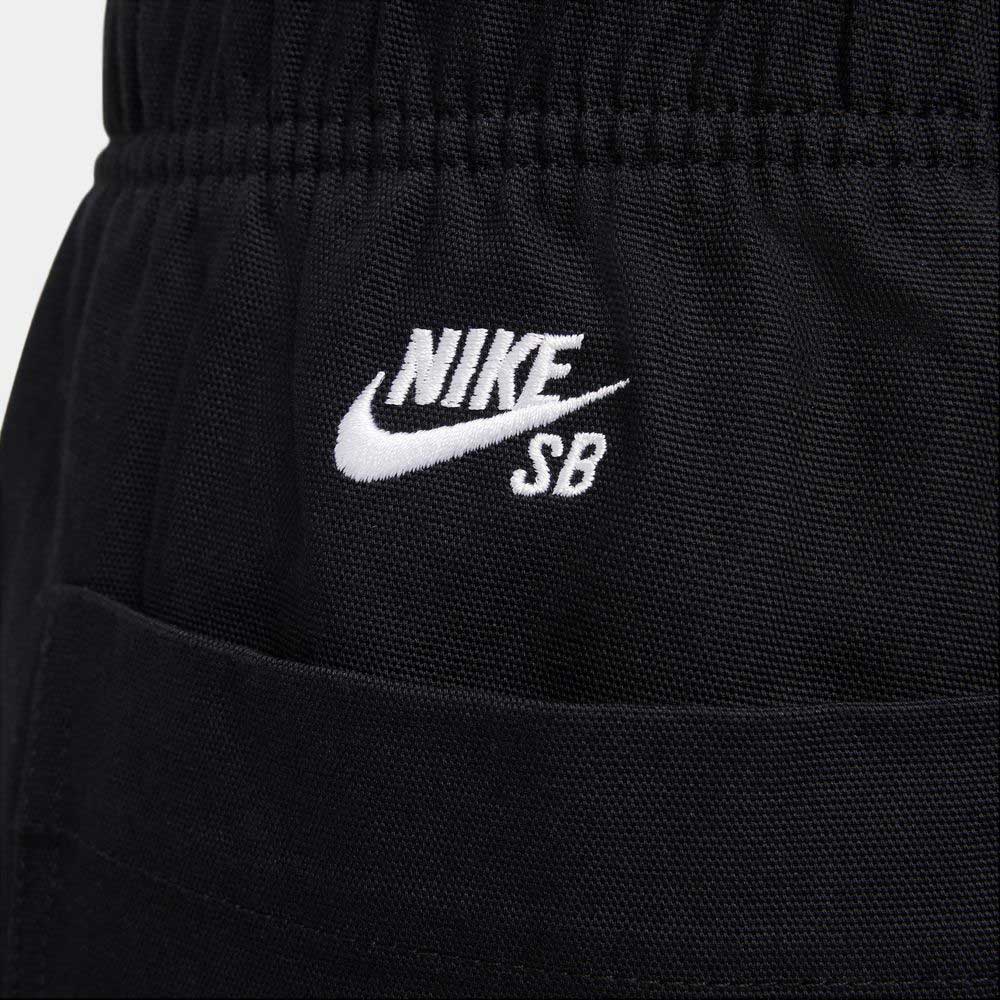 Close-up of the FV7349-010 Double Knee Pant Black by Nike SB Skateboarding, featuring a white embroidered NIKE SB logo with swoosh above the skate pants pocket seam on black cotton twill.