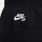 Close-up of the FV7349-010 Double Knee Pant Black by Nike SB Skateboarding, featuring a white embroidered NIKE SB logo with swoosh above the skate pants pocket seam on black cotton twill.