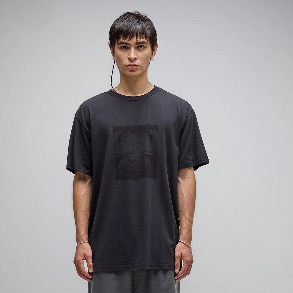 A young man with straight brown hair stands against a plain gray background, wearing the Crux Overdye Former Tee in Washed Black—a loose, vintage-inspired T-shirt crafted from 200GSM cotton with a subtle screen print graphic—paired with gray pants, looking directly at the camera with a neutral expression.