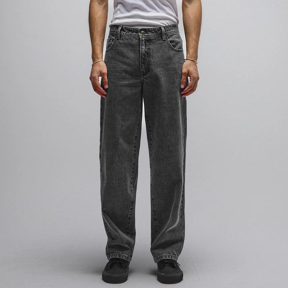A person stands facing forward against a plain light background, wearing a white t-shirt and the Crux Denim Pant Washed Black Former—loose-fitting, straight-fit jeans crafted from 100% cotton denim for comfort and durability. The person’s head is not visible.