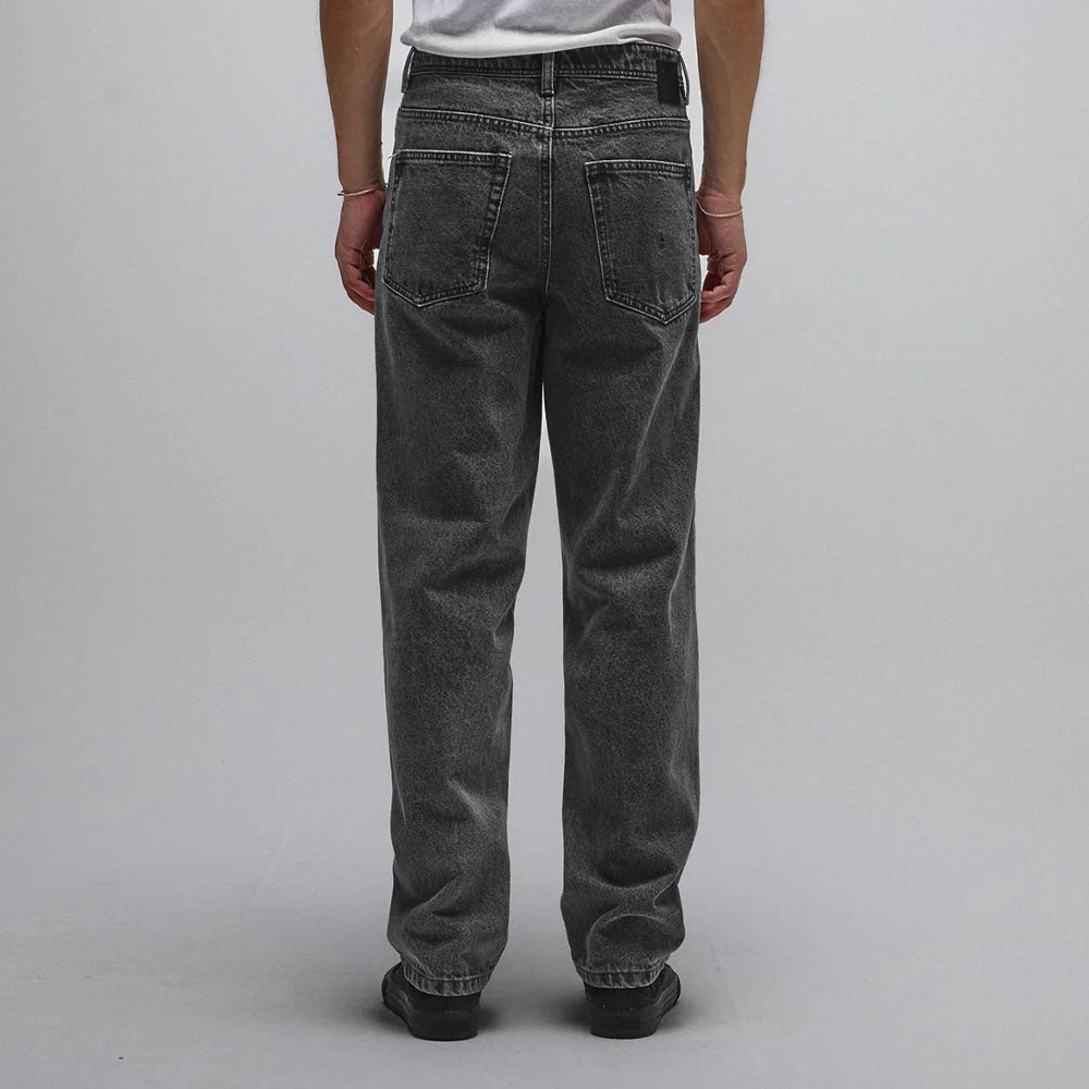 A person wearing a white t-shirt and loose-fitting, straight fit Crux Denim Pant Washed Black Former made from 100% cotton denim stands facing away from the camera against a plain light gray background.