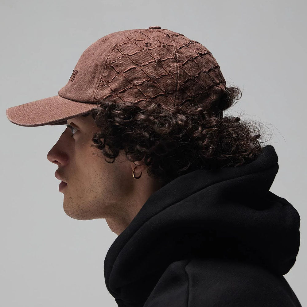 A person with curly hair wearing the Alleviate Cap Former Hat in Bark, a black hoodie layered over a vintage T-shirt, and a small hoop earring, shown in profile against a plain gray background.