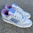 A pair of Forum 84 Low Adidas Skateboarding Gray Lavender Ice Blue Leather Shoes sits on rough concrete, featuring blue stripes, purple lining, and Adidas branding on the tongue.