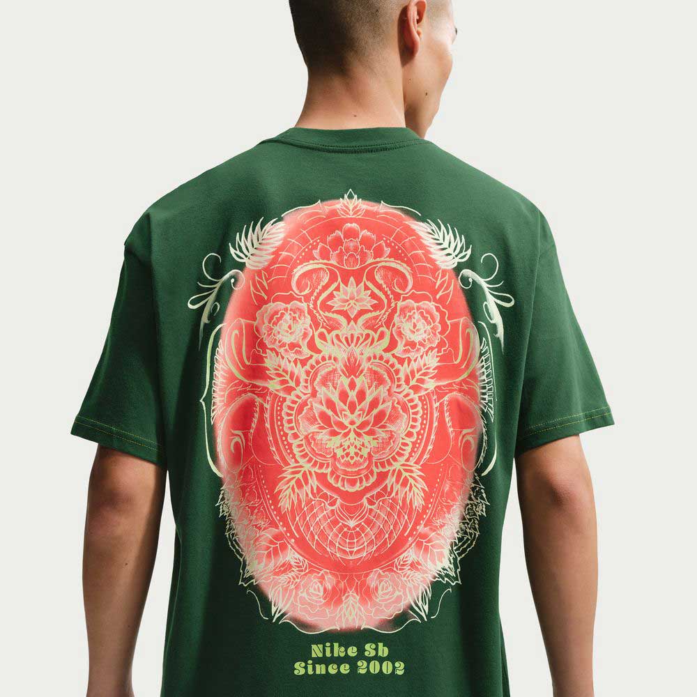 Someone wears the Gorge Green Som Tum Dunk Tee IH1478341, a roomy Nike SB Skateboarding T-shirt in dark green midweight cotton with a large red and white floral mandala on the back and "Nike Sb Since 2002" in yellow at the lower back.