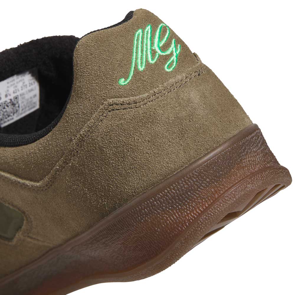Close-up of the heel of a Gray Black Mark Gonzales KJ0281 Aloha Super Suede Skateboarding Shoe in olive green suede, featuring ‘Mg’ embroidered in green on the back. The textured brown sole adds contrast, while a portion of the inner label is visible.
